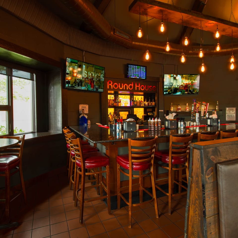 Warmly lit Round House restaurant bar with red-upholstered stools, pendant lights, televisions, and bottles displayed behind the counter.