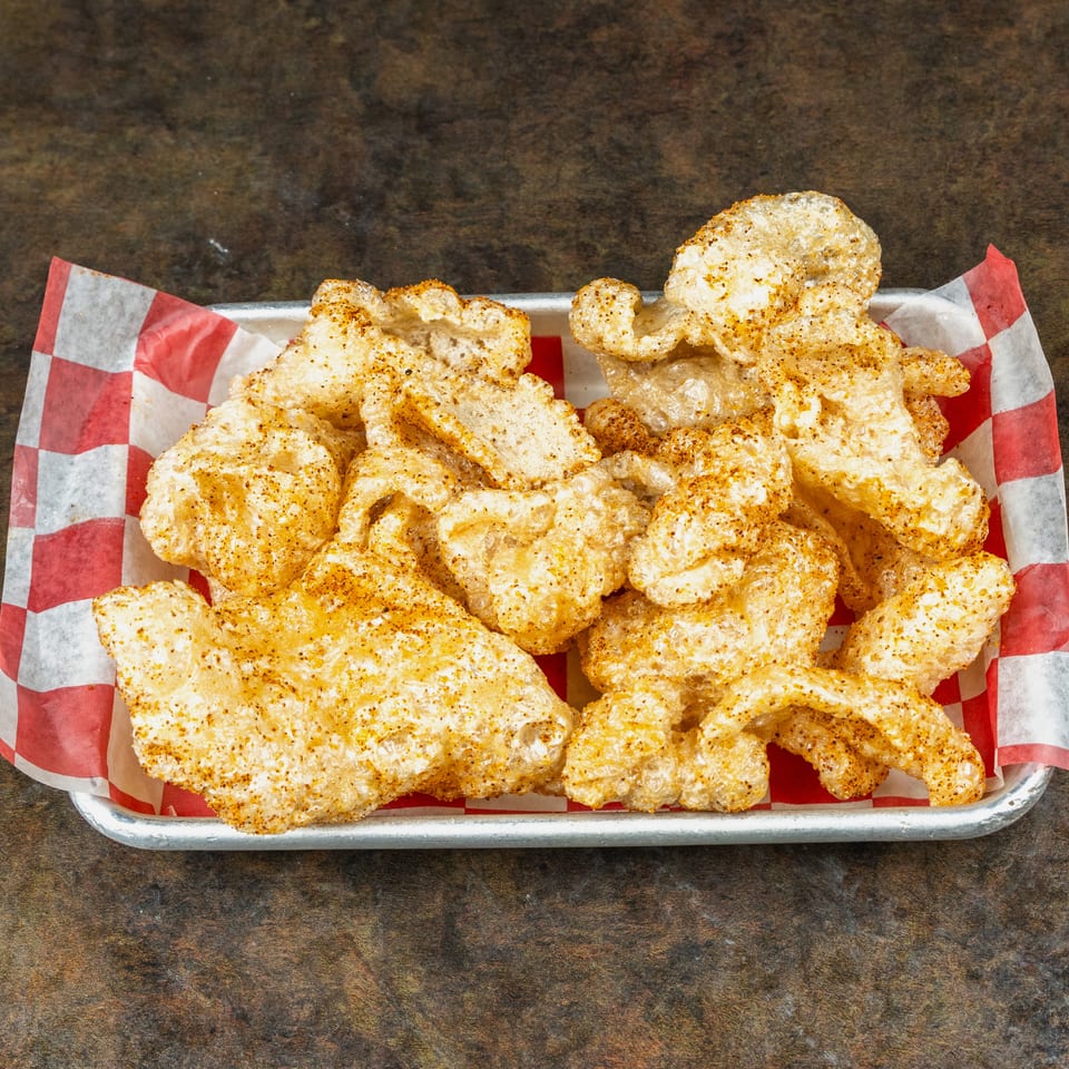 Seasoned pork rinds piled in a metal tray lined with red-and-white checkered paper on a wooden table.