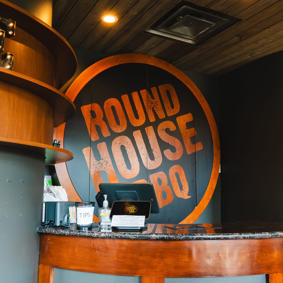 Restaurant host counter with illuminated marquee letters, wood accents, and a large Round House BBQ logo.