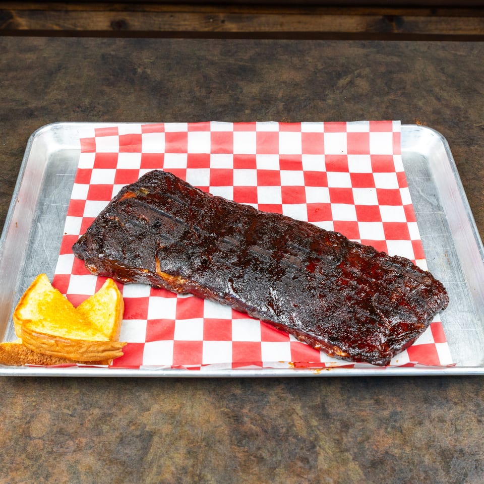 Glazed barbecue ribs with toasted bread on a red-and-white checkered paper-lined metal tray.