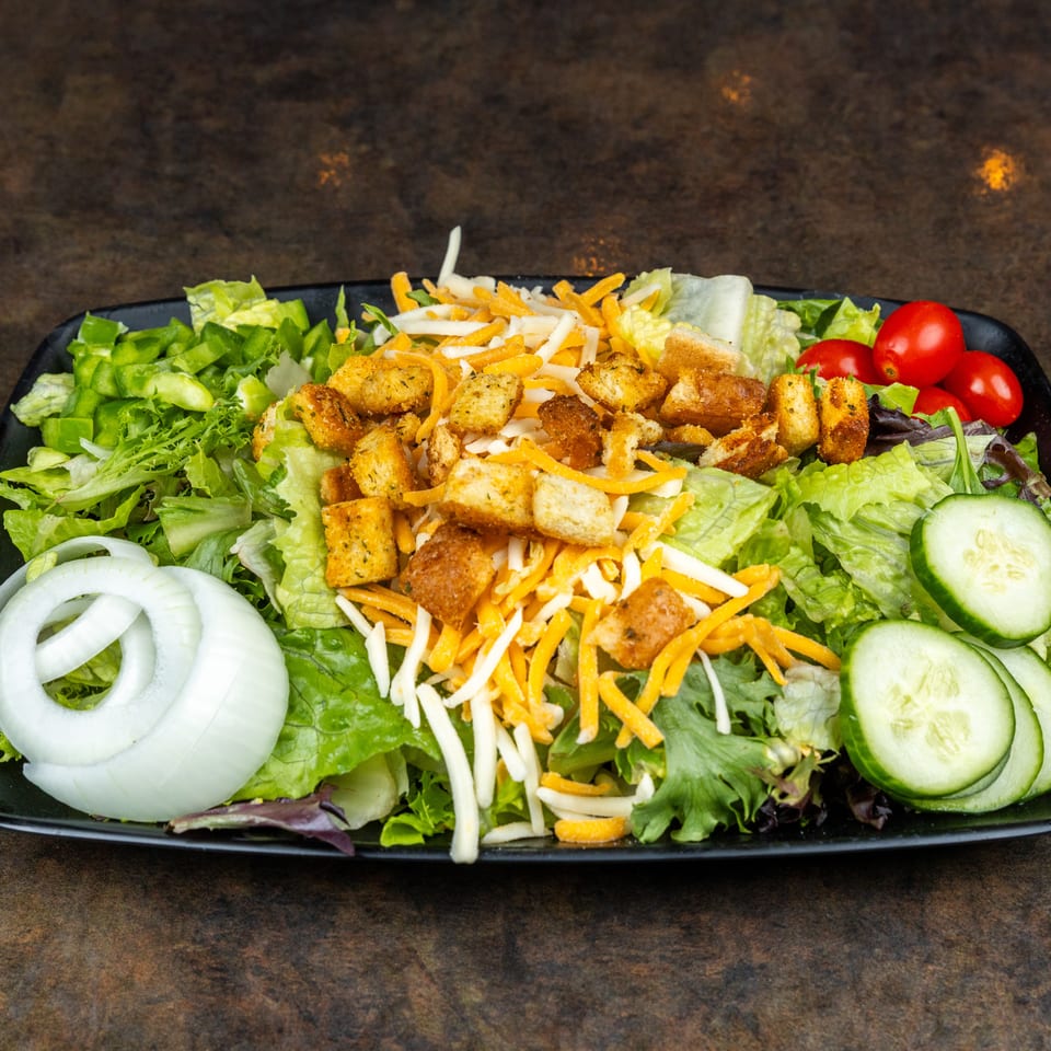 Fresh garden salad topped with shredded cheddar cheese, croutons, lettuce, tomatoes, cucumbers, and onion rings on a black platter.