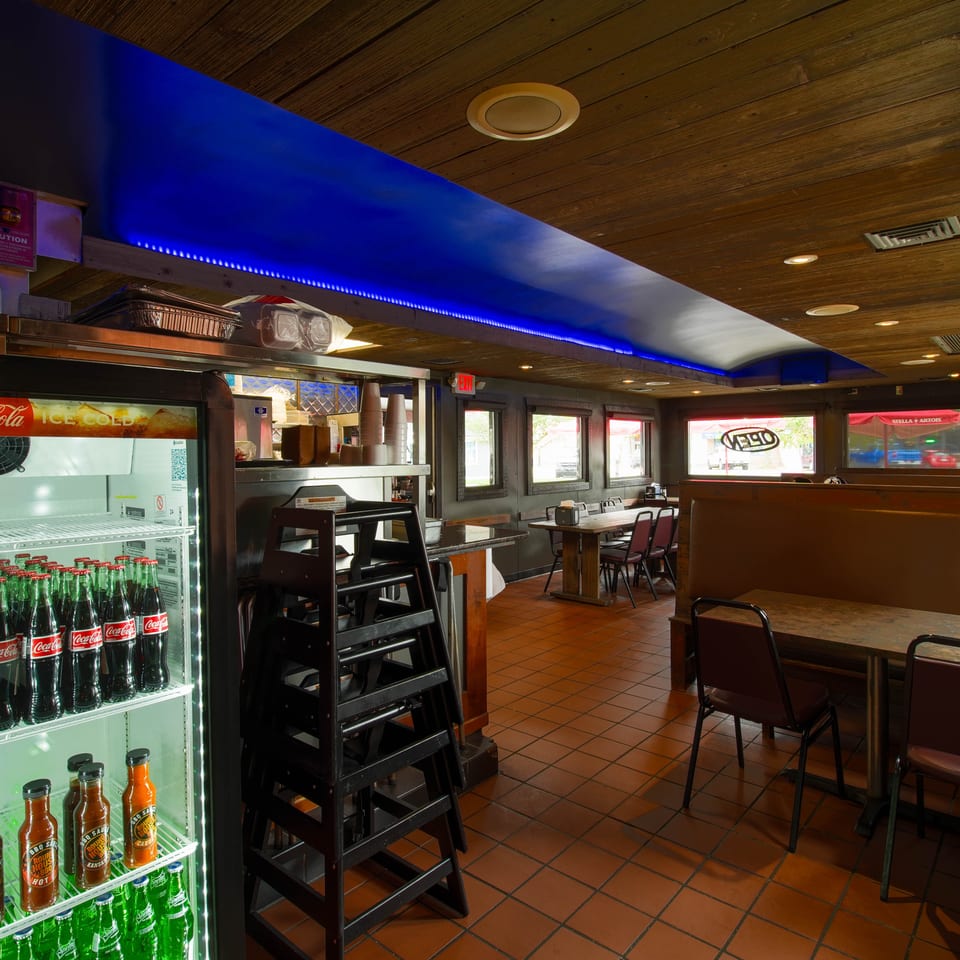 Restaurant interior with blue accent lighting, wood-paneled ceiling, tiled floors, booths, tables, and a Coca-Cola beverage cooler.