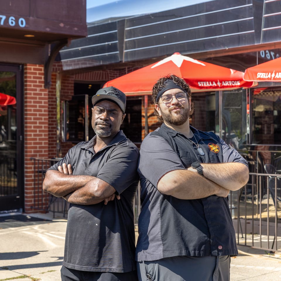Two restaurant staff members pose with crossed arms outside a brick building beneath red Stella Artois patio umbrellas.