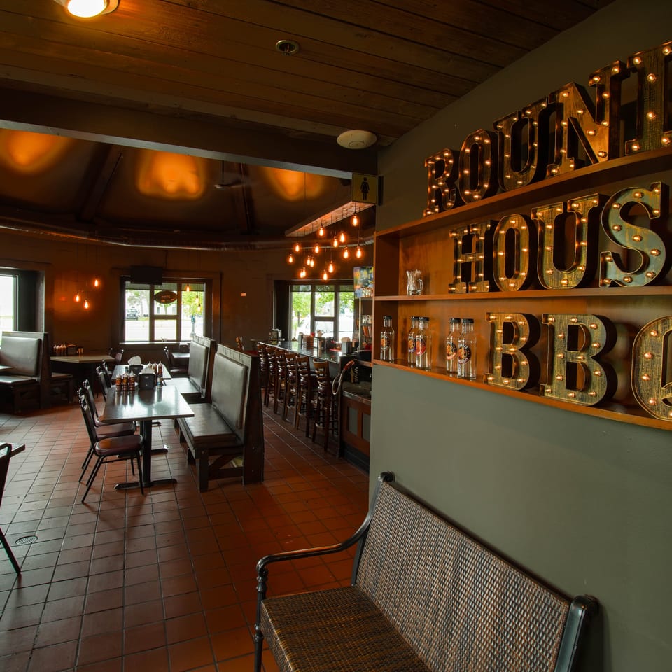 Warmly lit restaurant interior with booths, tables, pendant lights, and illuminated “ROU... HOUSE BBQ” marquee letters.