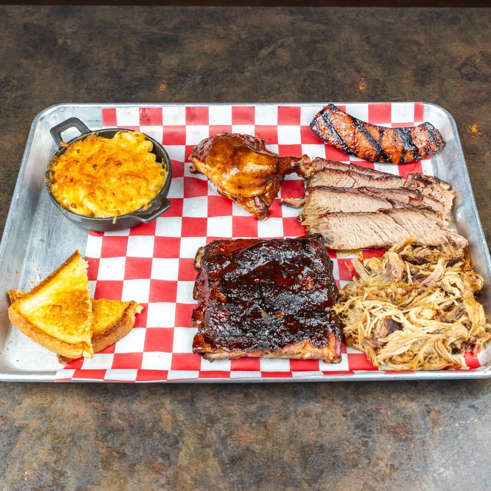 Barbecue platter with ribs, sliced brisket, pulled pork, chicken, sausage, macaroni and cheese, and toast on checkered paper.